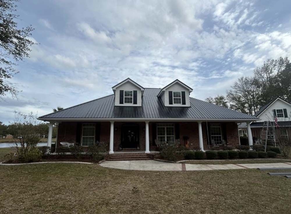 Brick house with dark metal roof, dormers, and porch under a cloudy sky.