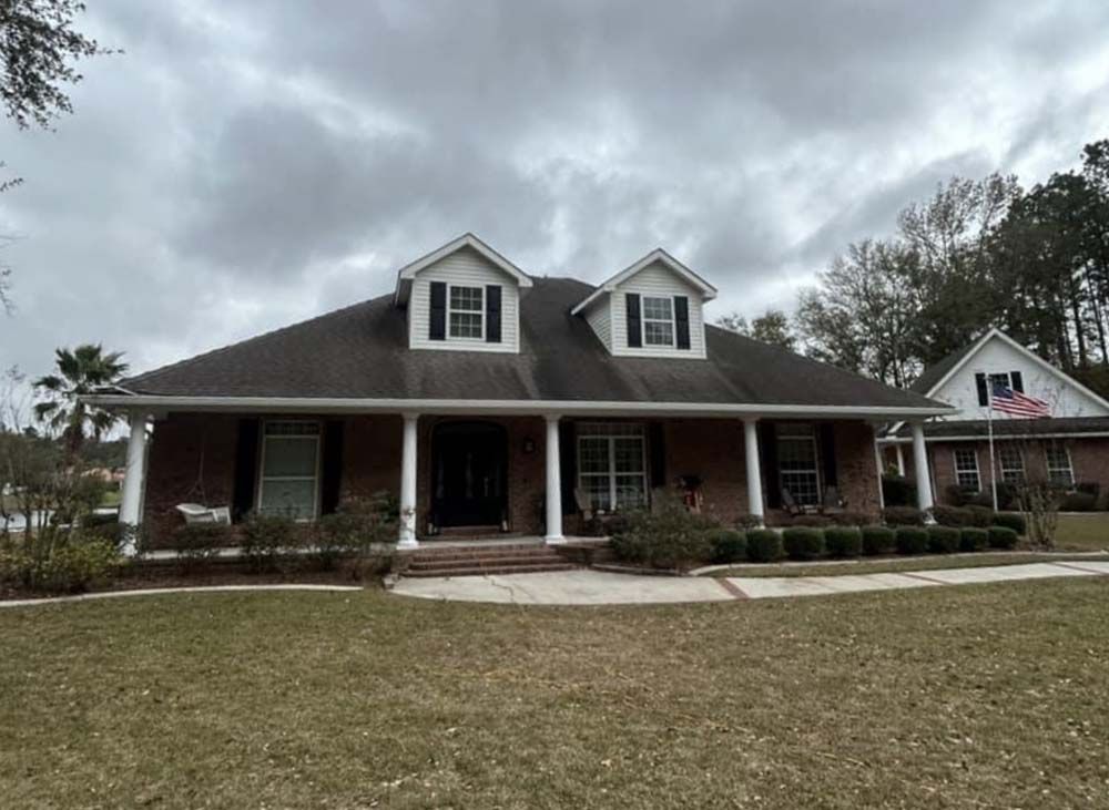 Brick house with a long porch, dormers, and black shutters under a cloudy sky.