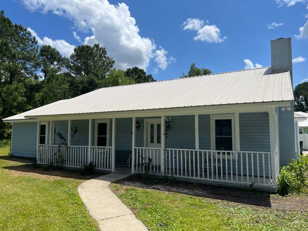 Blue house with white porch, metal roof, and chimney, under a blue sky with clouds.