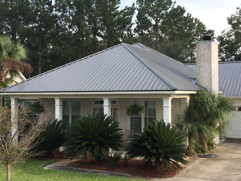 House with a gray metal roof, white siding, and front porch. Palm trees and greenery in front.