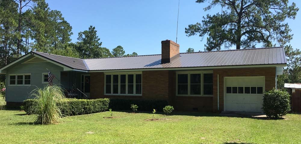 A single-story brick house with a metal roof, chimney, and garage door on a sunny day.