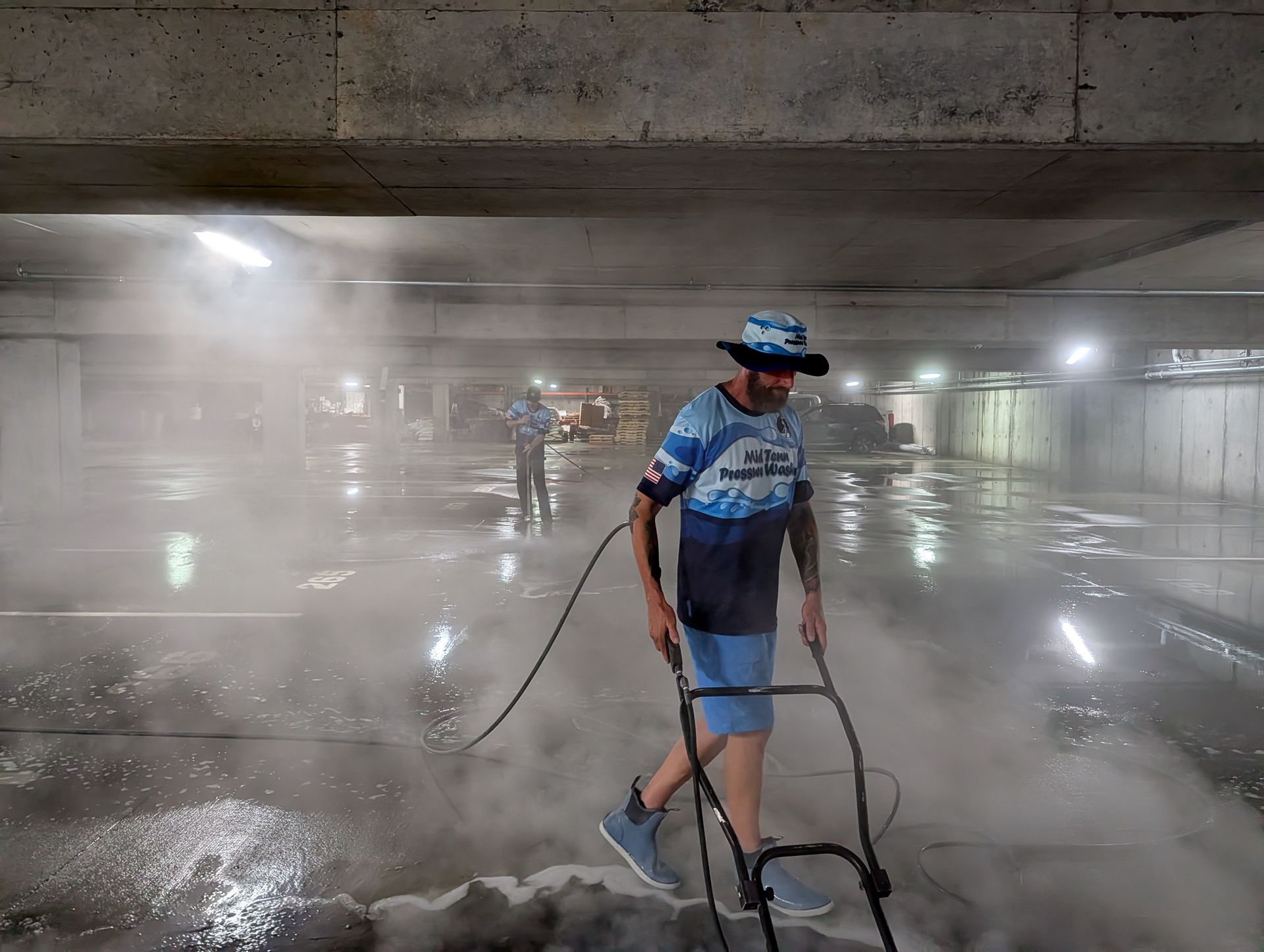 Worker pressure-washing a concrete parking garage floor with heavy mist and reflections