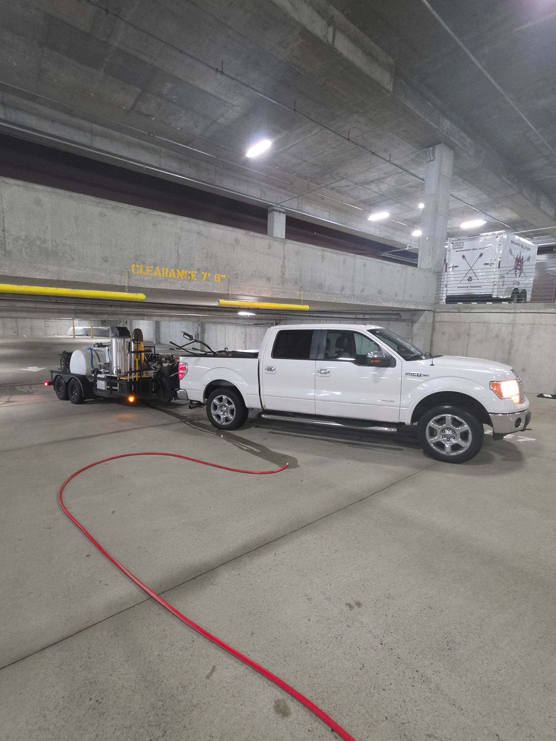 White pickup truck with tow equipment parked in a concrete garage, with a red hose on the floor.
