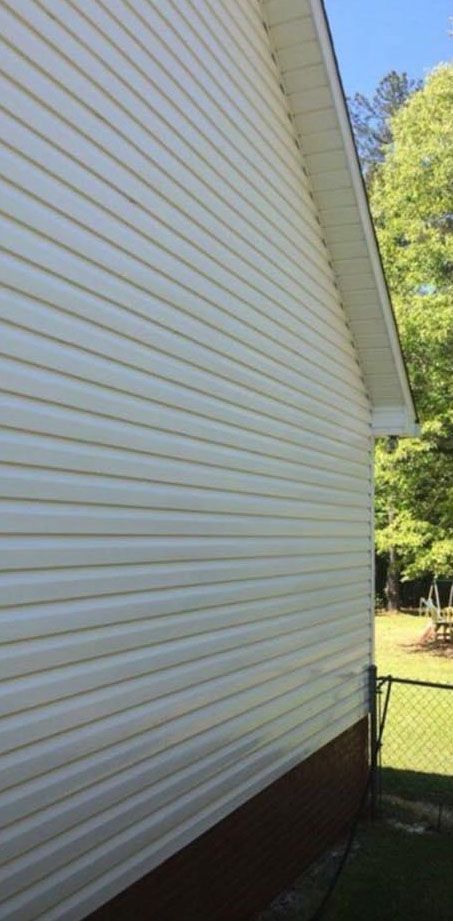 White vinyl siding on a house with brown trim at the bottom, a chain-link fence, and trees.