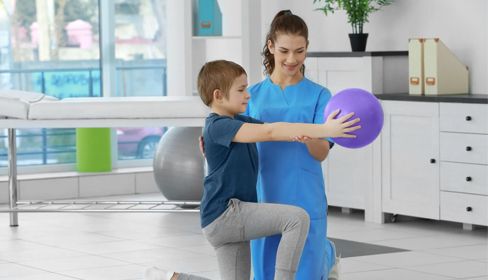 A child in gray pants kneels, holding a purple ball with help from a healthcare provider in a blue uniform.