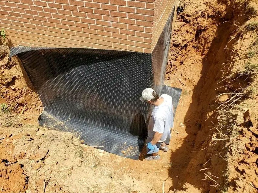 A construction worker applies a black dimpled drainage membrane to a brick foundation wall in an excavated trench.