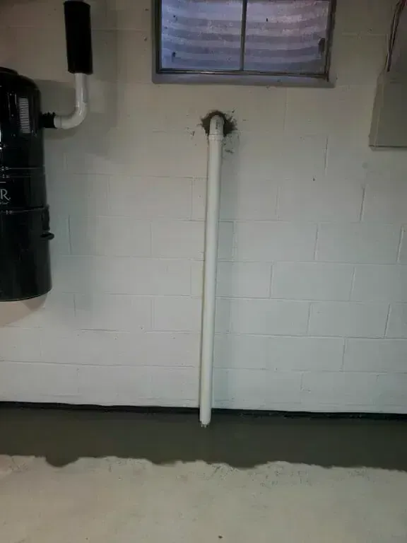 A white PVC pipe runs vertically down a painted white concrete block wall, anchored through the wall near a basement window.