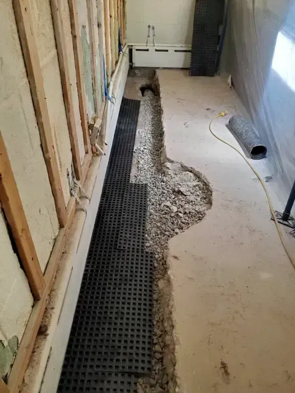 An interior basement floor with a trench cut along a wooden-framed wall, showing black dimpled drainage mat and gravel.