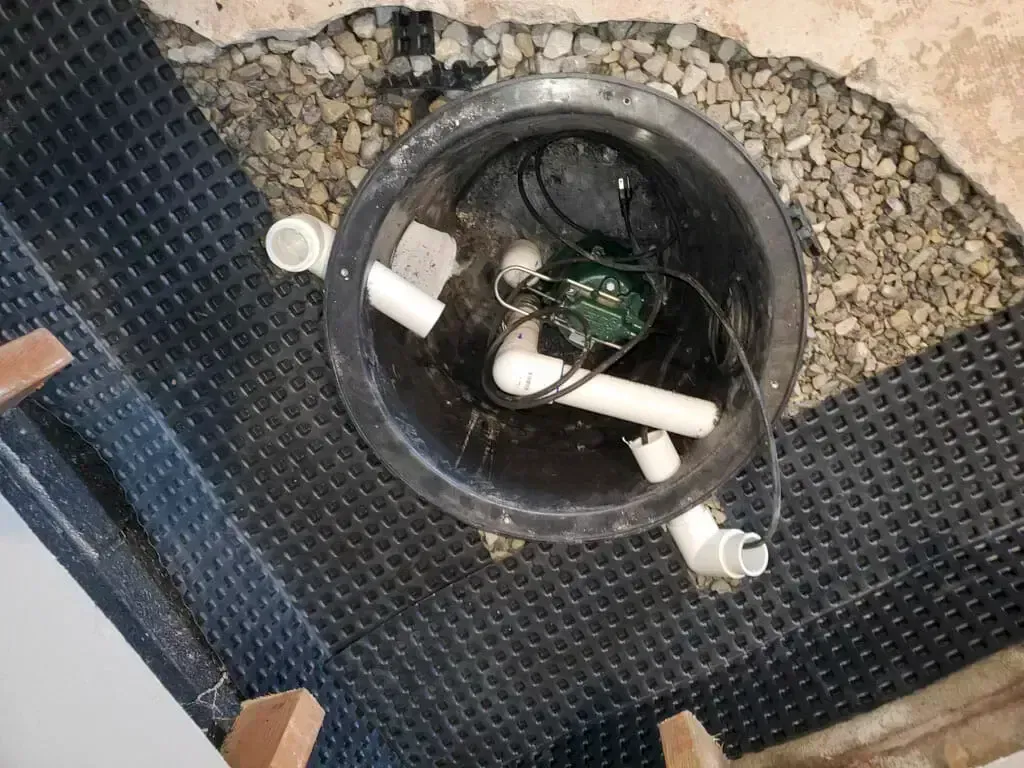 A black sump pump basin set in gravel with white PVC inlet and discharge pipes and an electrical pump inside.