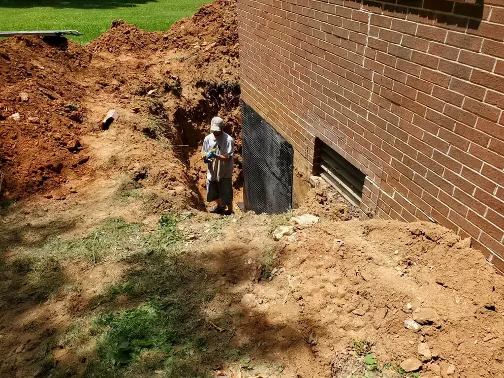A person works in a deep trench dug along the brick foundation of a house, where waterproof coating is visible on the wall.