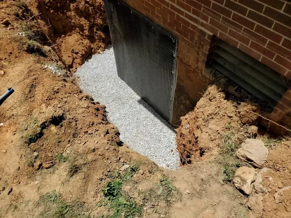 A black foundation drainage membrane installed against a brick wall, surrounded by gravel fill in an excavated trench.