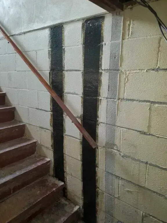 Two black carbon fiber reinforcement strips installed vertically on a beige concrete block wall next to a wooden staircase.