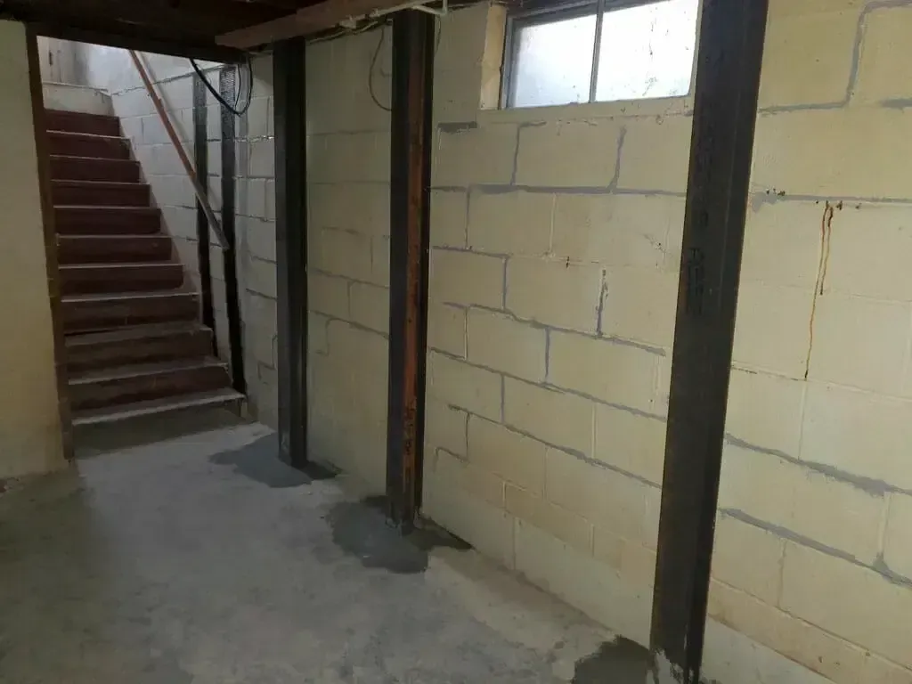 A basement interior with a wooden staircase and concrete block walls reinforced by vertical steel support beams.