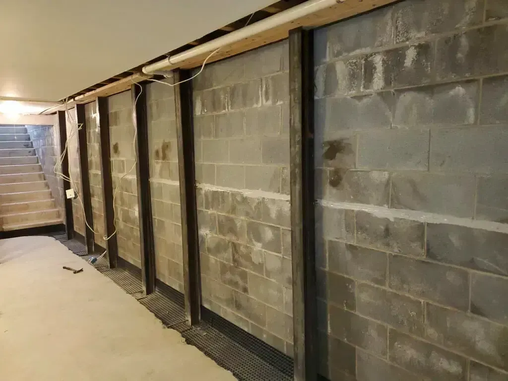 A basement wall reinforced with several vertical steel I-beams, with stairs visible in the background.