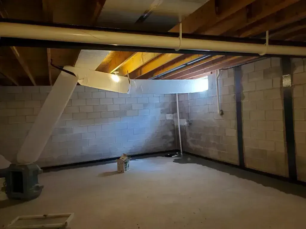 A basement with exposed wooden floor joists, concrete block walls featuring structural supports, and white ductwork.