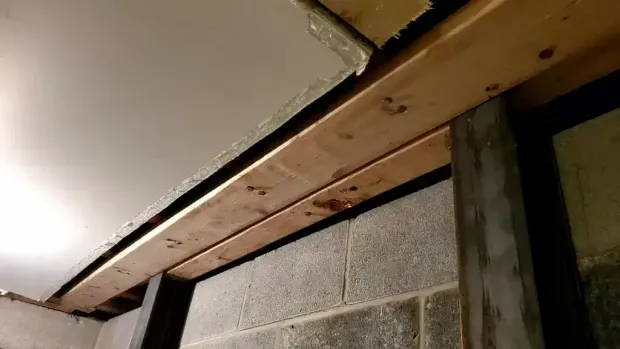 A low-angle view of a wooden beam installed horizontally against a grey concrete block wall in an unfinished basement.