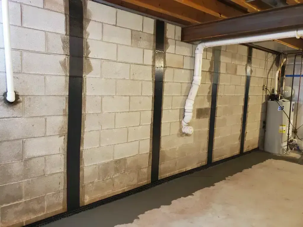 Basement cinder block wall reinforced with vertical black carbon fiber straps above a floor-level drainage system.