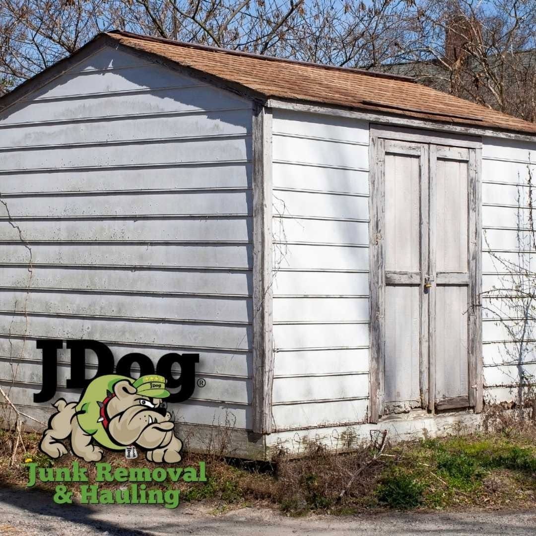 Small white shed with double doors and a tan roof in a grassy yard, with JDog Junk Removal & Hauling logo in the corner