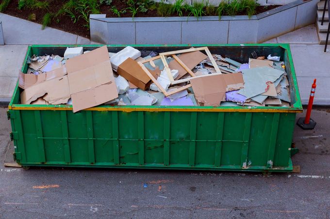 Green dumpster filled with broken cardboard and debris beside a curb