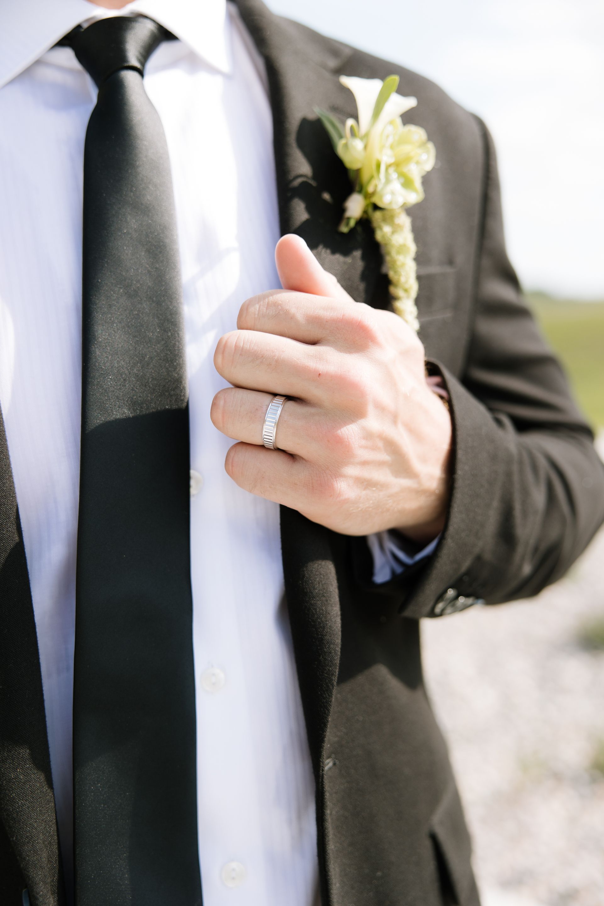 Man in black suit, white shirt, and tie, with boutonniere; hand on jacket showing ring.