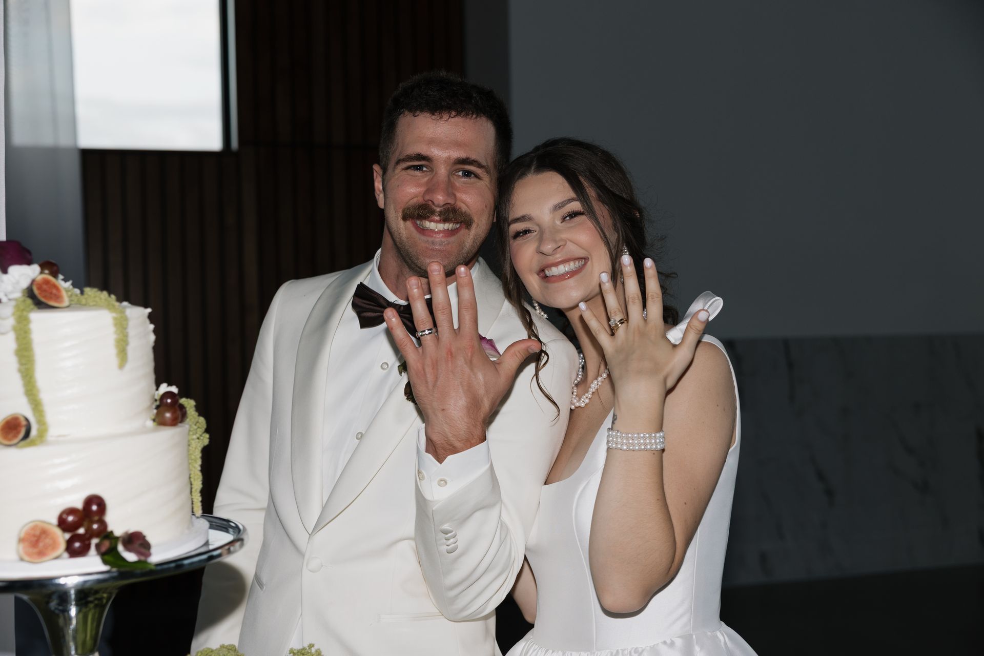 Newlyweds smiling, showing off rings near wedding cake. Man in white suit, woman in white dress.