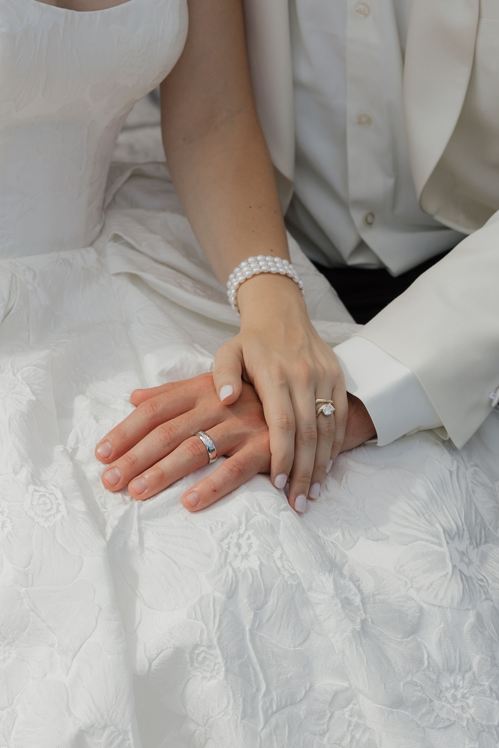 Two hands, each with a wedding ring, clasped together over white fabric. One wears a bracelet.