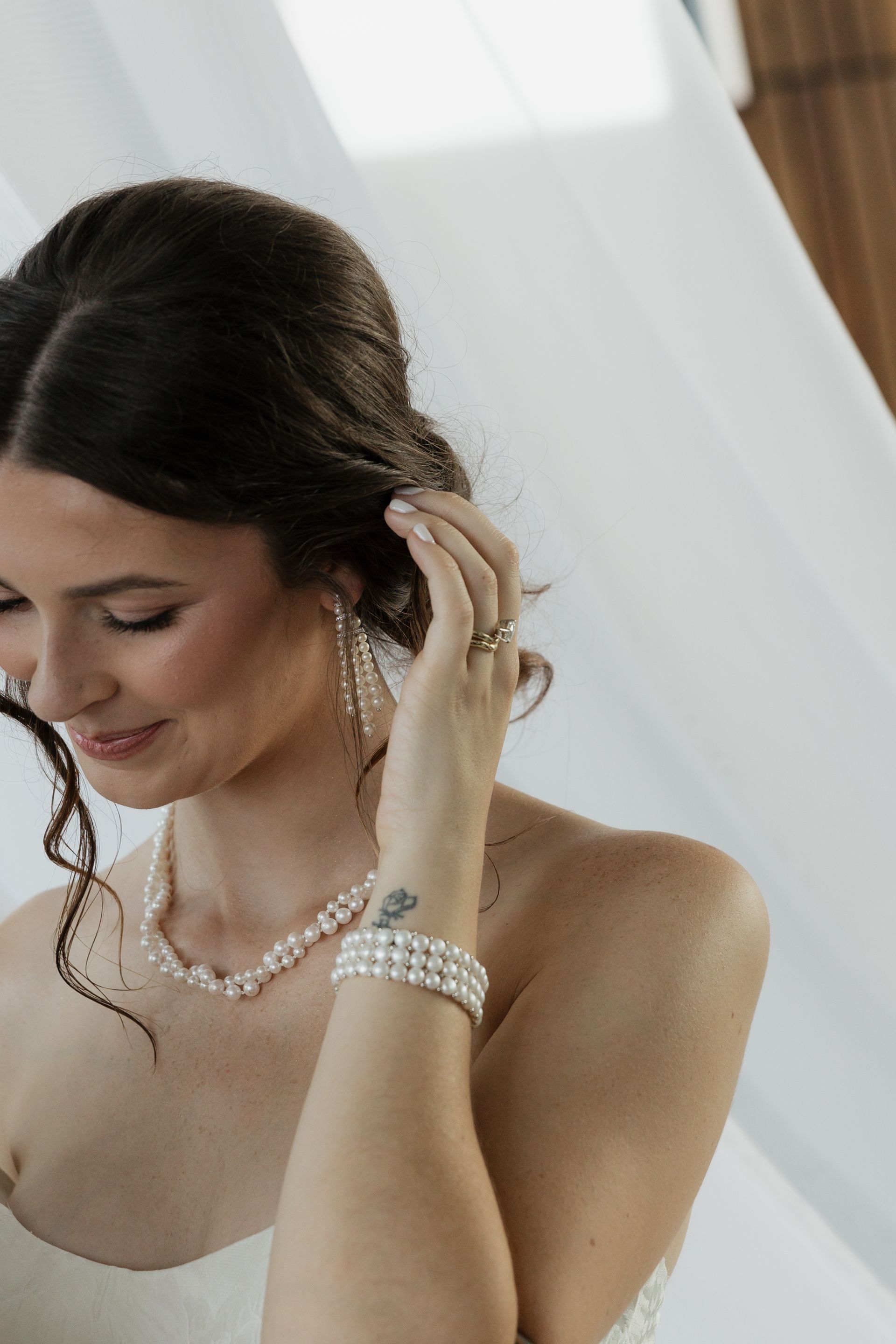 Bride in a strapless gown, adjusting her hair. She wears pearl jewelry and has a tattoo on her wrist.