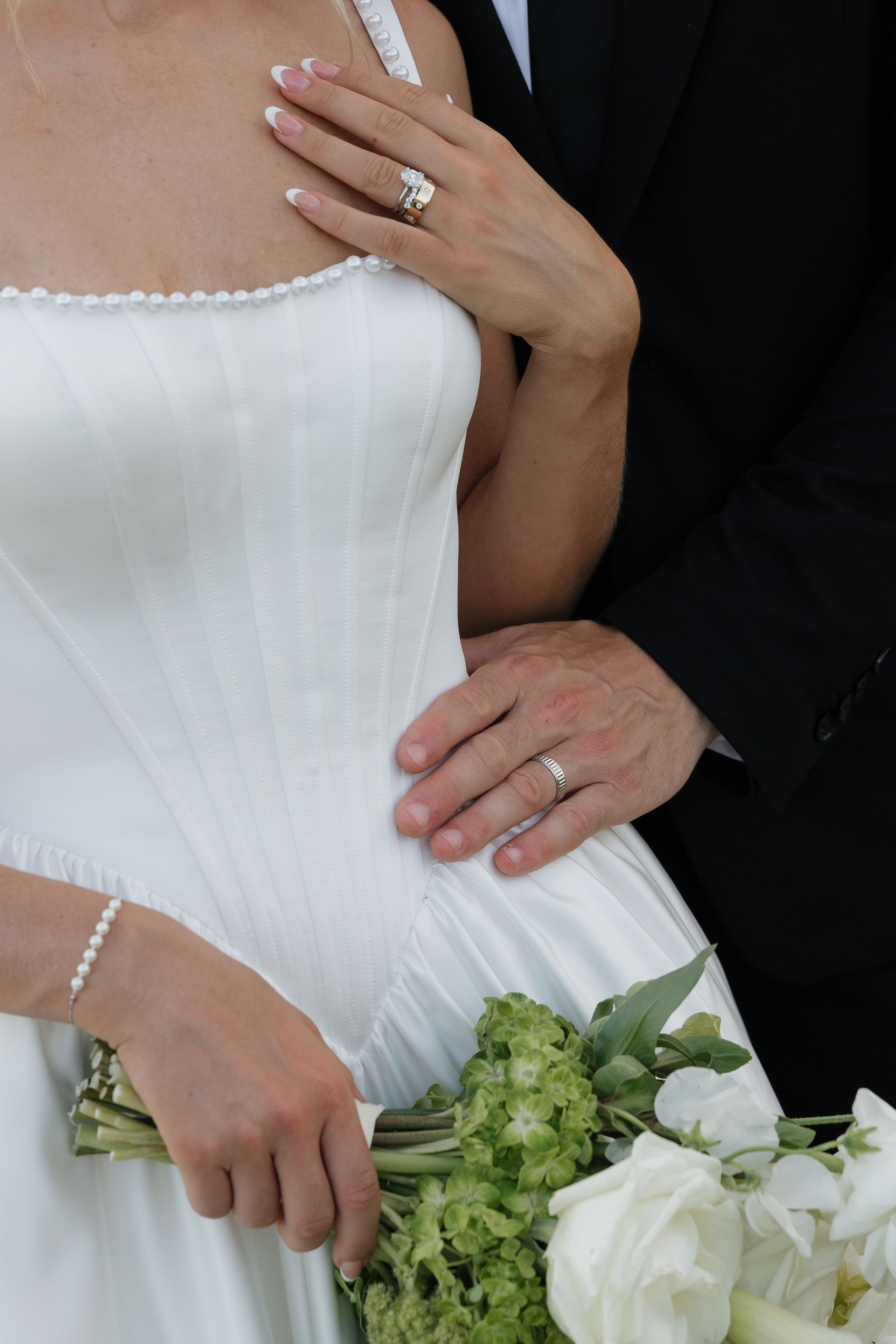 Bride in white gown, hand on chest, bouquet. Groom's hand on waist, both with wedding rings.