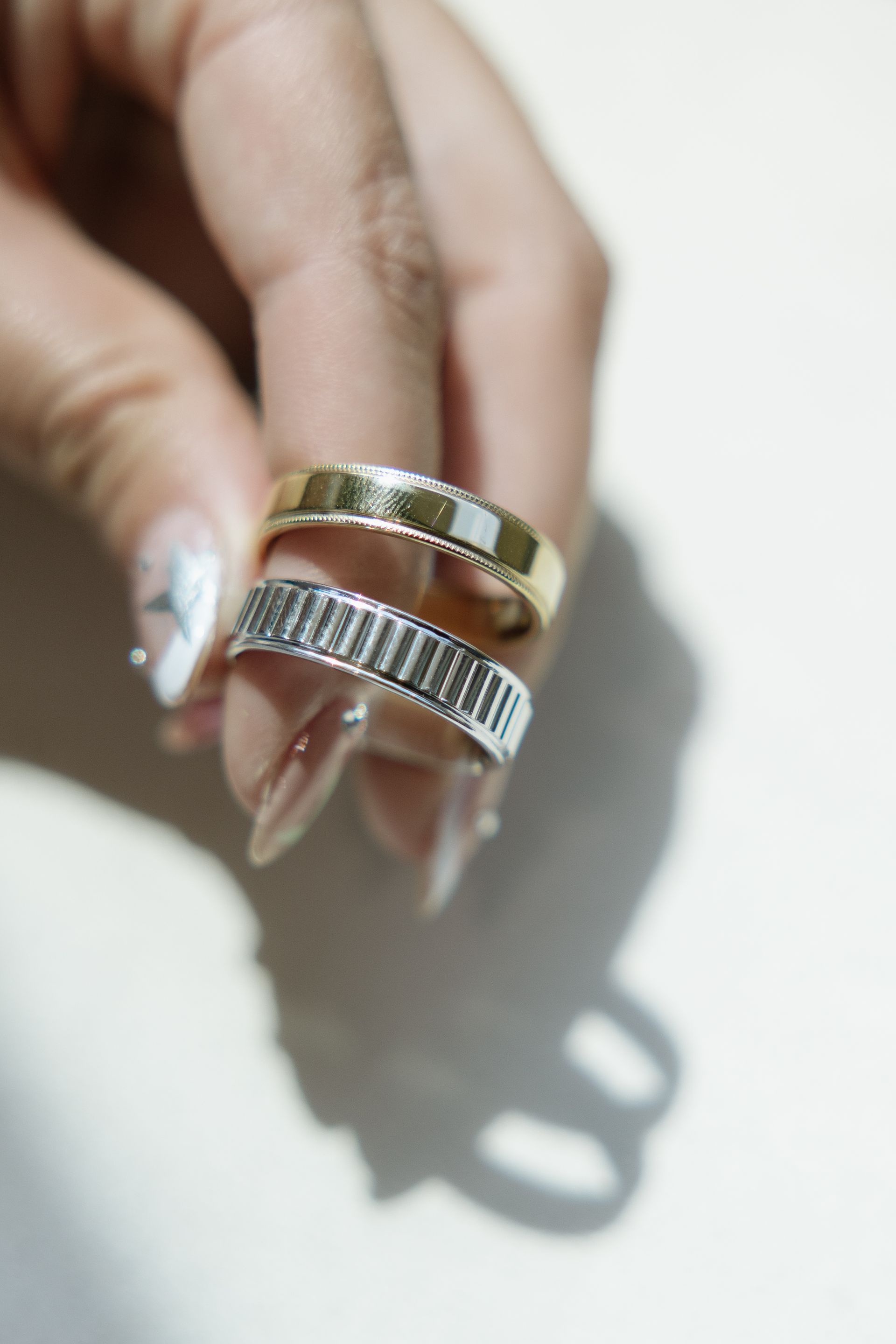 Hand holding two patterned rings, one gold, one silver, on a white surface.