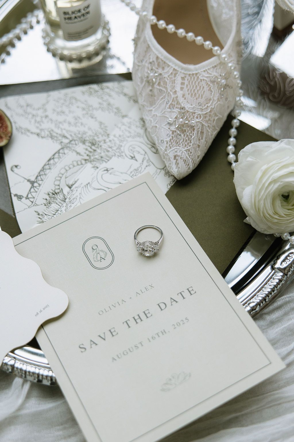 Wedding details: Save the date, engagement ring, lace shoe, pearl necklace, and white flower on a tray.