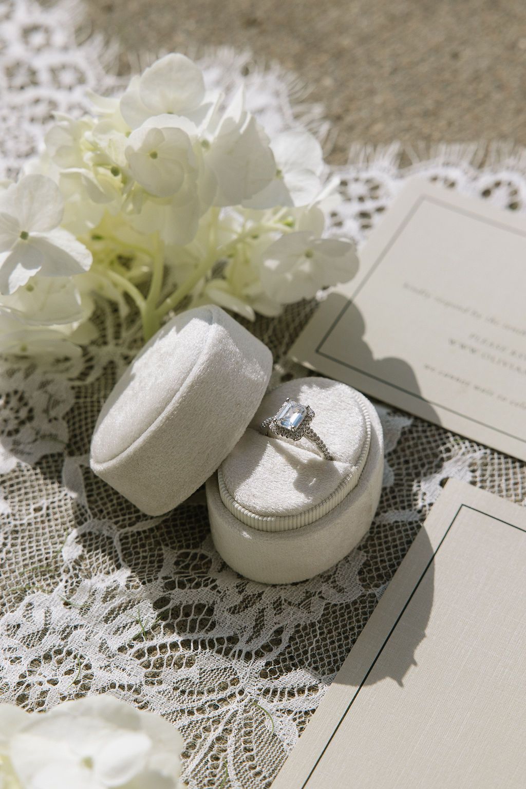 Engagement ring in a velvet box, with white flowers and invitation on lace and burlap.