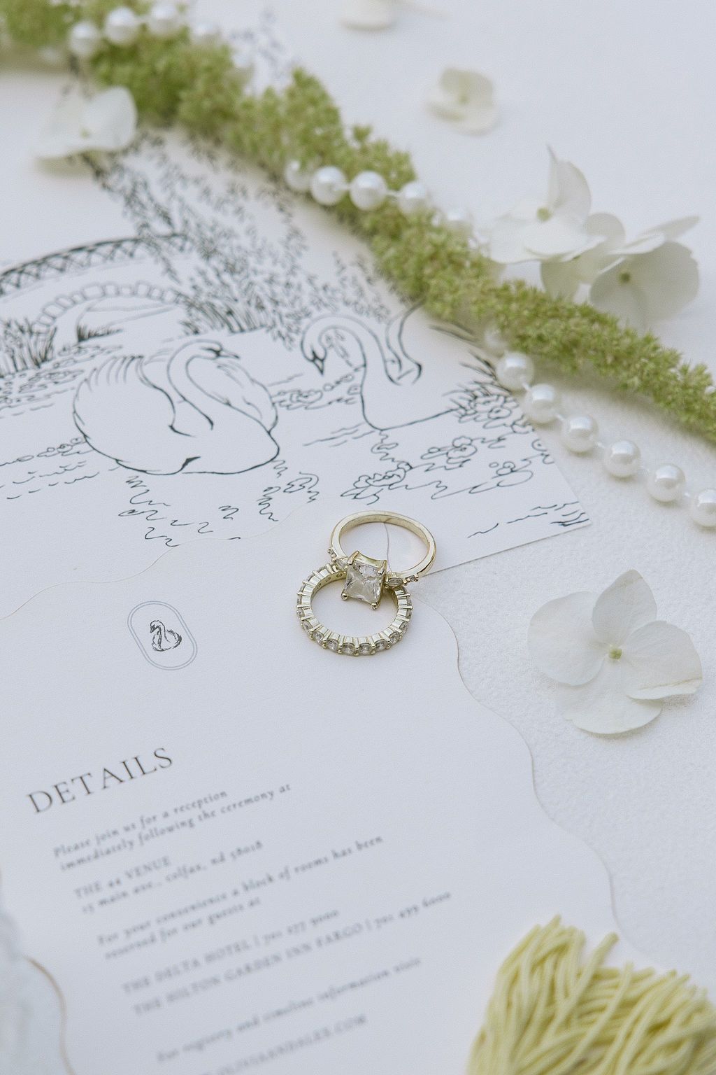 Wedding rings on invitation with swan illustration, surrounded by flowers, pearls, and greenery.