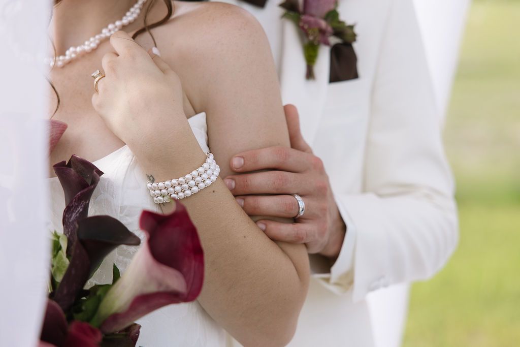 Bride and groom, arms around each other, wedding rings visible. Bride wears white dress and pearls, holding flowers.