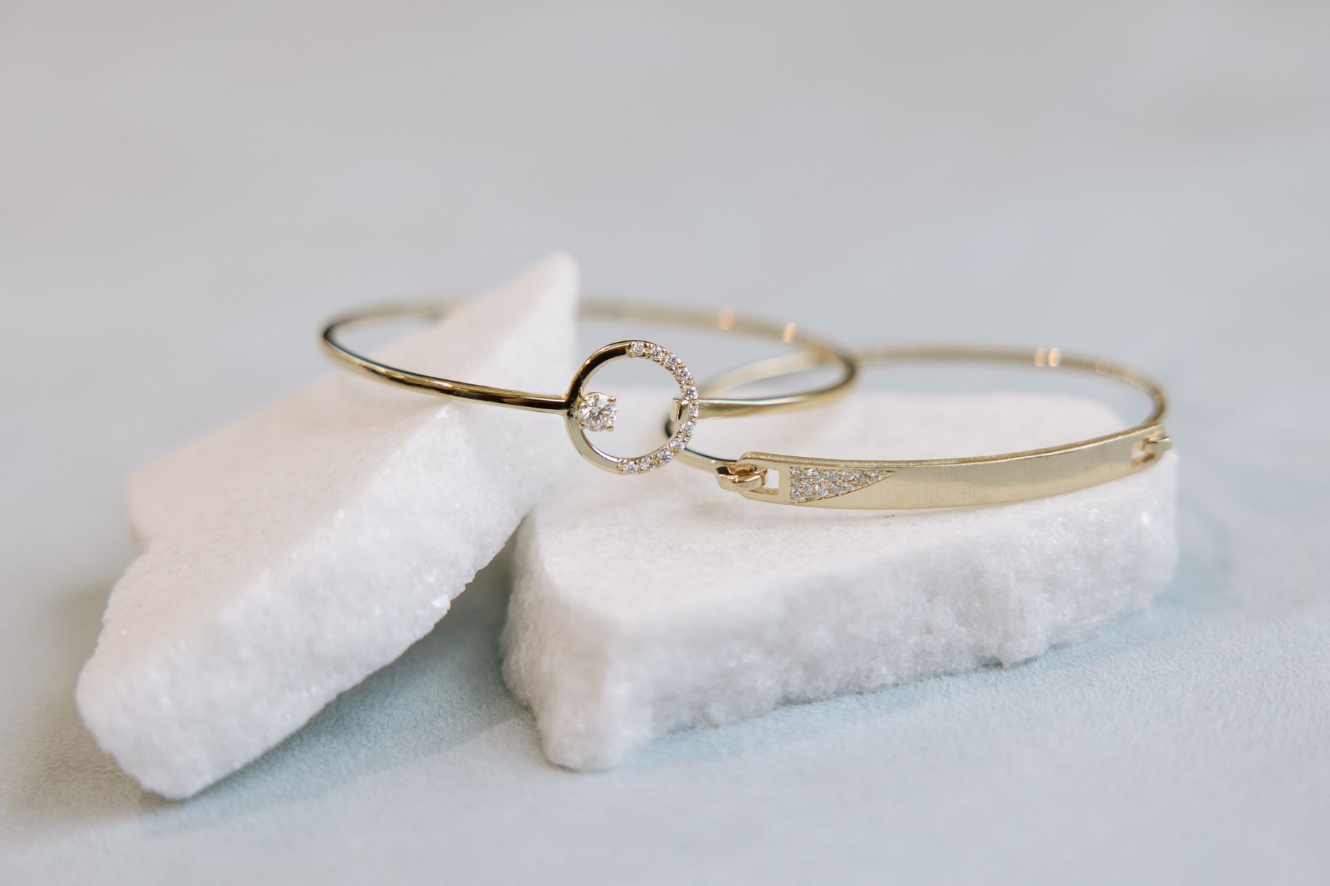 Two gold bracelets, one with a circle and gem, resting on white, textured blocks.