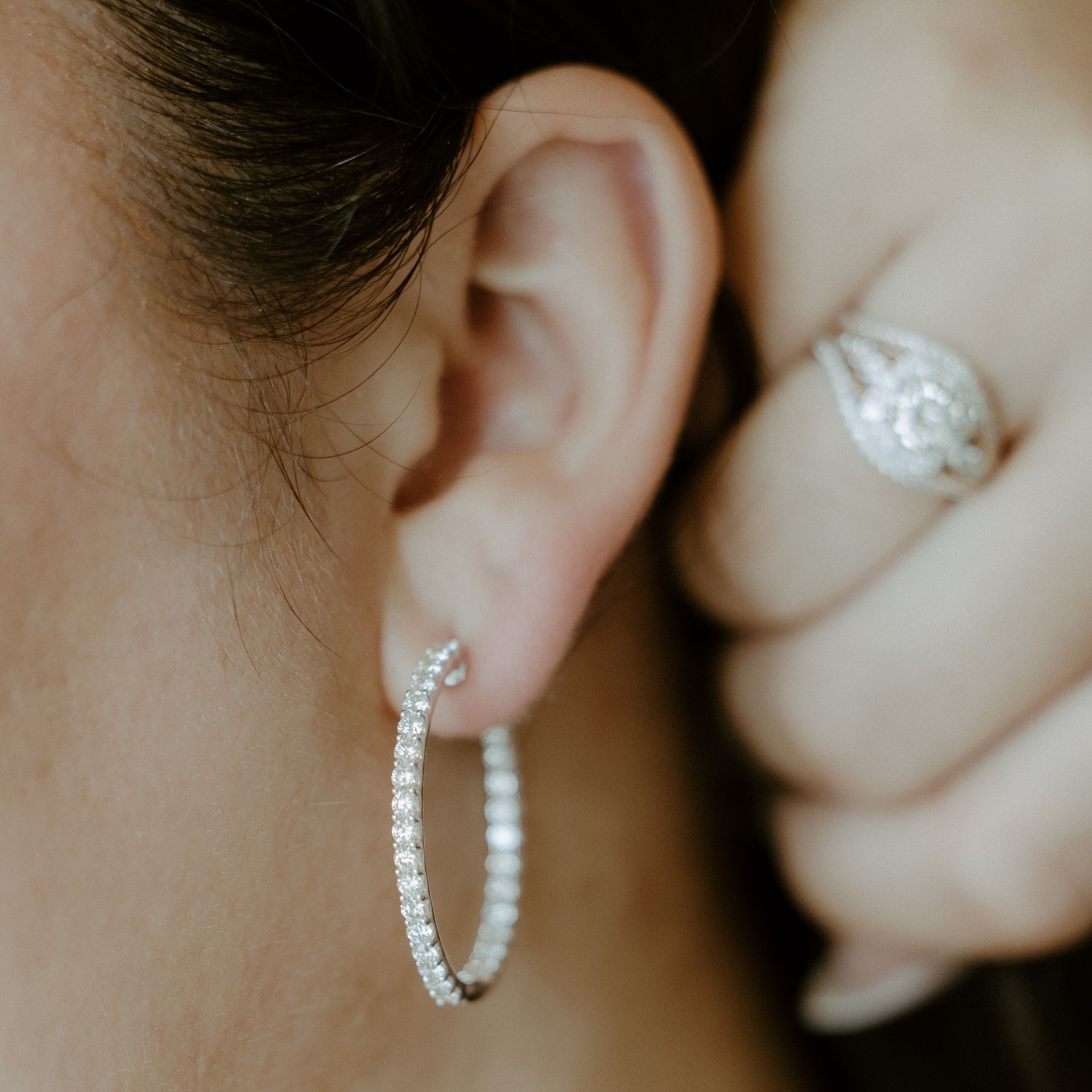 Woman's ear with diamond hoop earring; hand with diamond ring in the background.