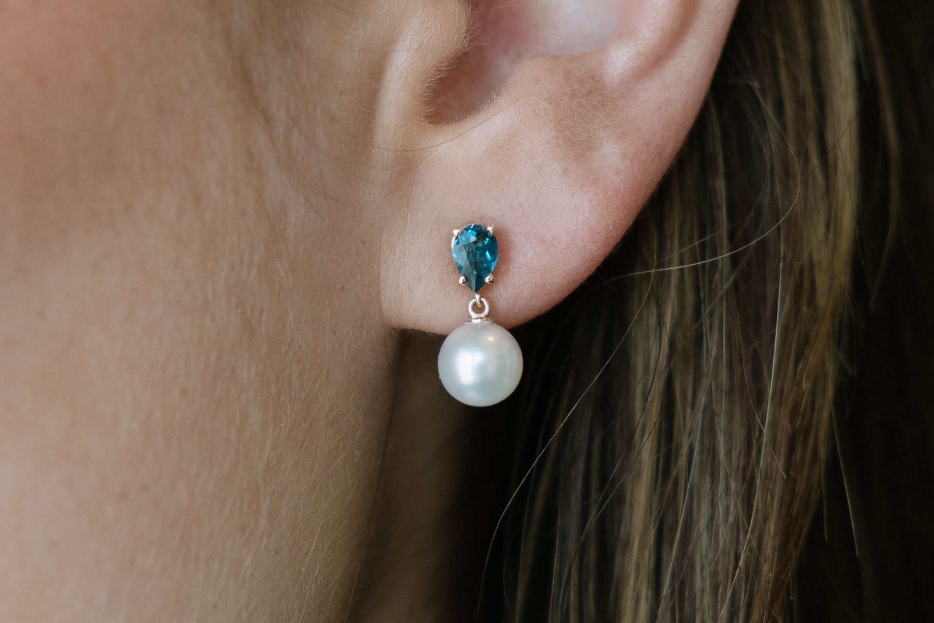 Earring with a blue gem and a white pearl against a person's ear.