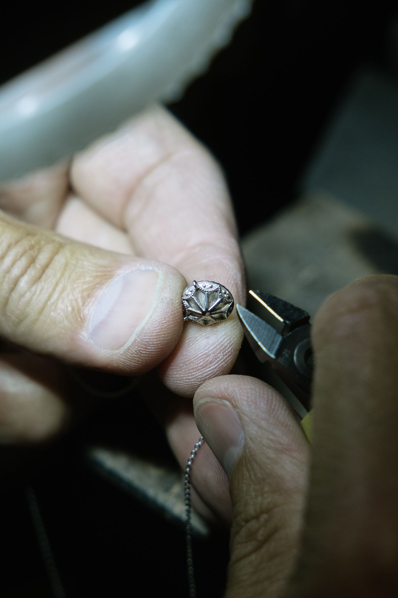 Hands holding a small gemstone with pliers and a chain in a dimly lit setting.