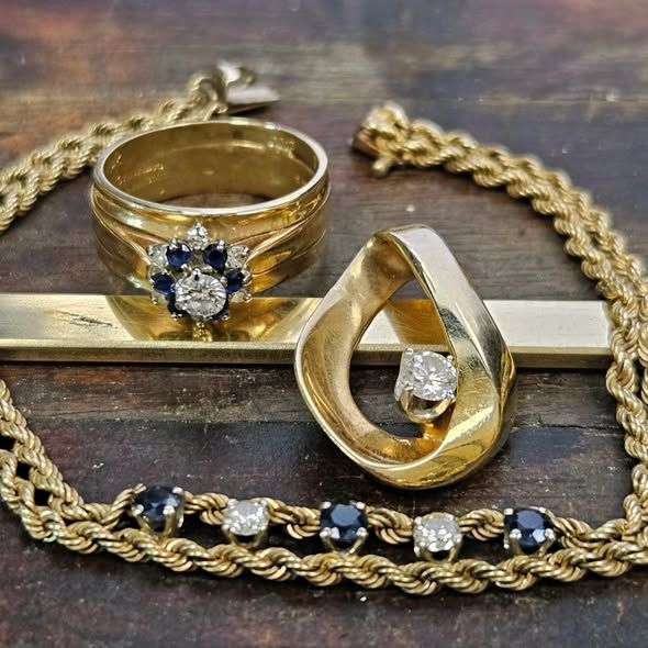 Gold jewelry: ring with gemstones, pendant with diamond, and bracelet with blue stones, on wooden surface.
