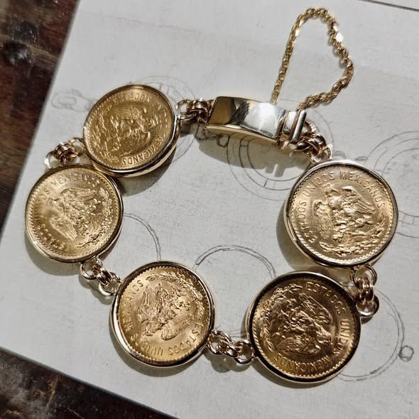 Gold coin bracelet with six Mexican coins linked by a gold chain, lying on a sketched paper.