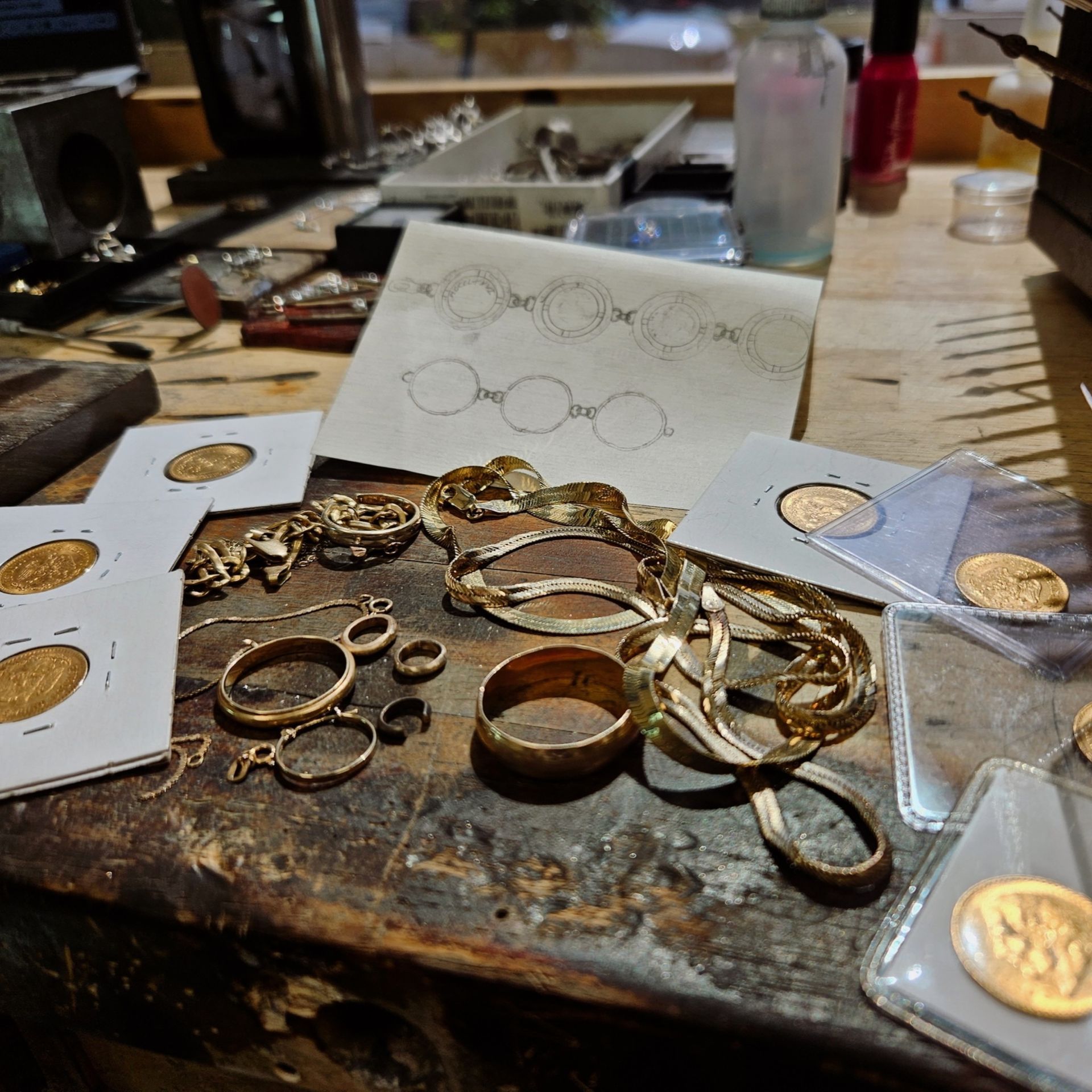 Jewelry pieces and gold coins on a workbench, with design sketch and tools, lit by natural light.