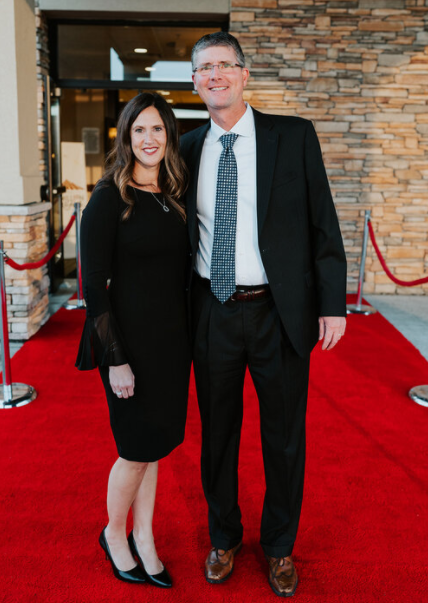 Couple posing on a red carpet. Woman in black dress, man in suit and tie. Building background.