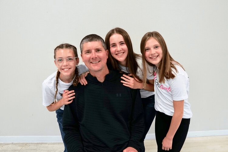 Man smiles with three girls, posing in front of a white background. Girls have arms around the man.
