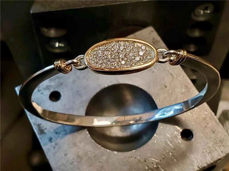 Silver and gold bracelet with diamond-encrusted oval center, resting on a metal tool.
