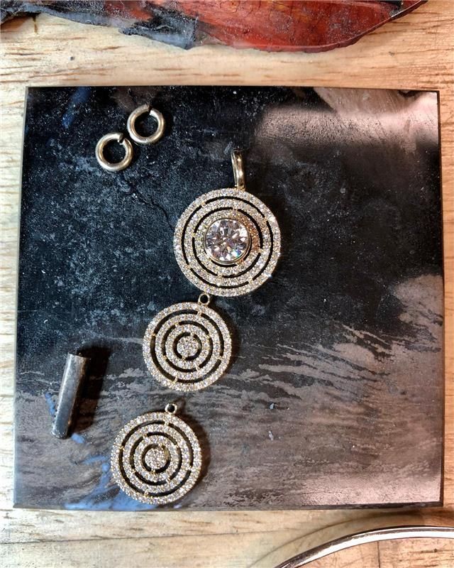 Jewelry pieces on a jeweler's bench. Three gold pendant circles with diamonds, two jump rings, and a metal tool.