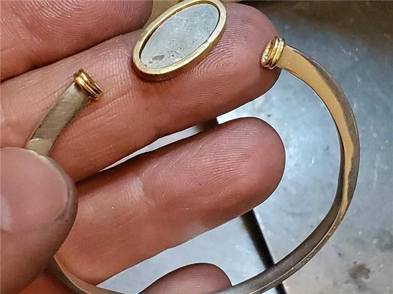 Close-up of a silver bracelet with gold accents and a detached oval element held by a hand.