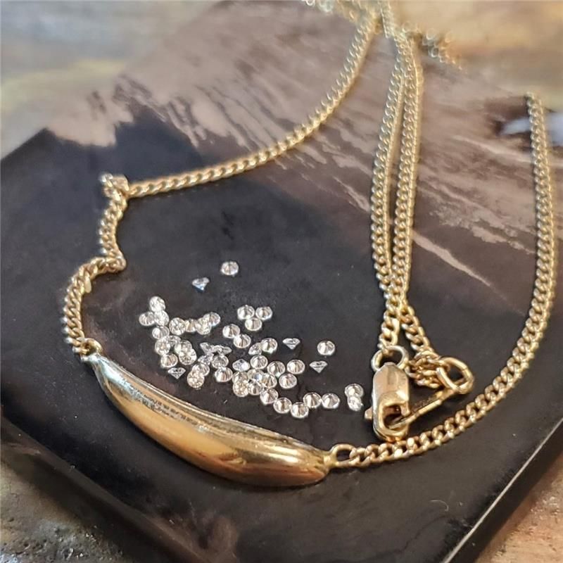 Gold necklace with a curved pendant, resting on a dark, textured surface with scattered small crystals.