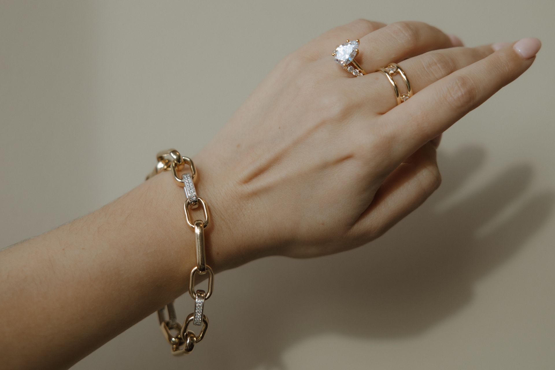 Hand wearing gold chain bracelet and two rings against a beige background.