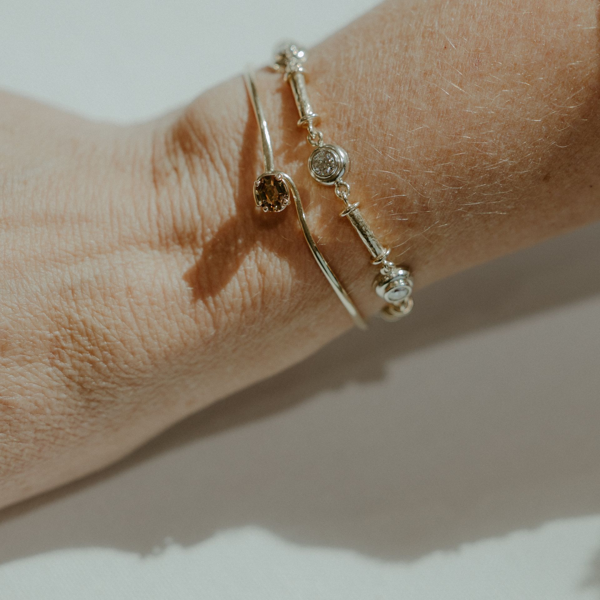 Wrist with two delicate gold bracelets, one with a brown stone, the other with clear stones.
