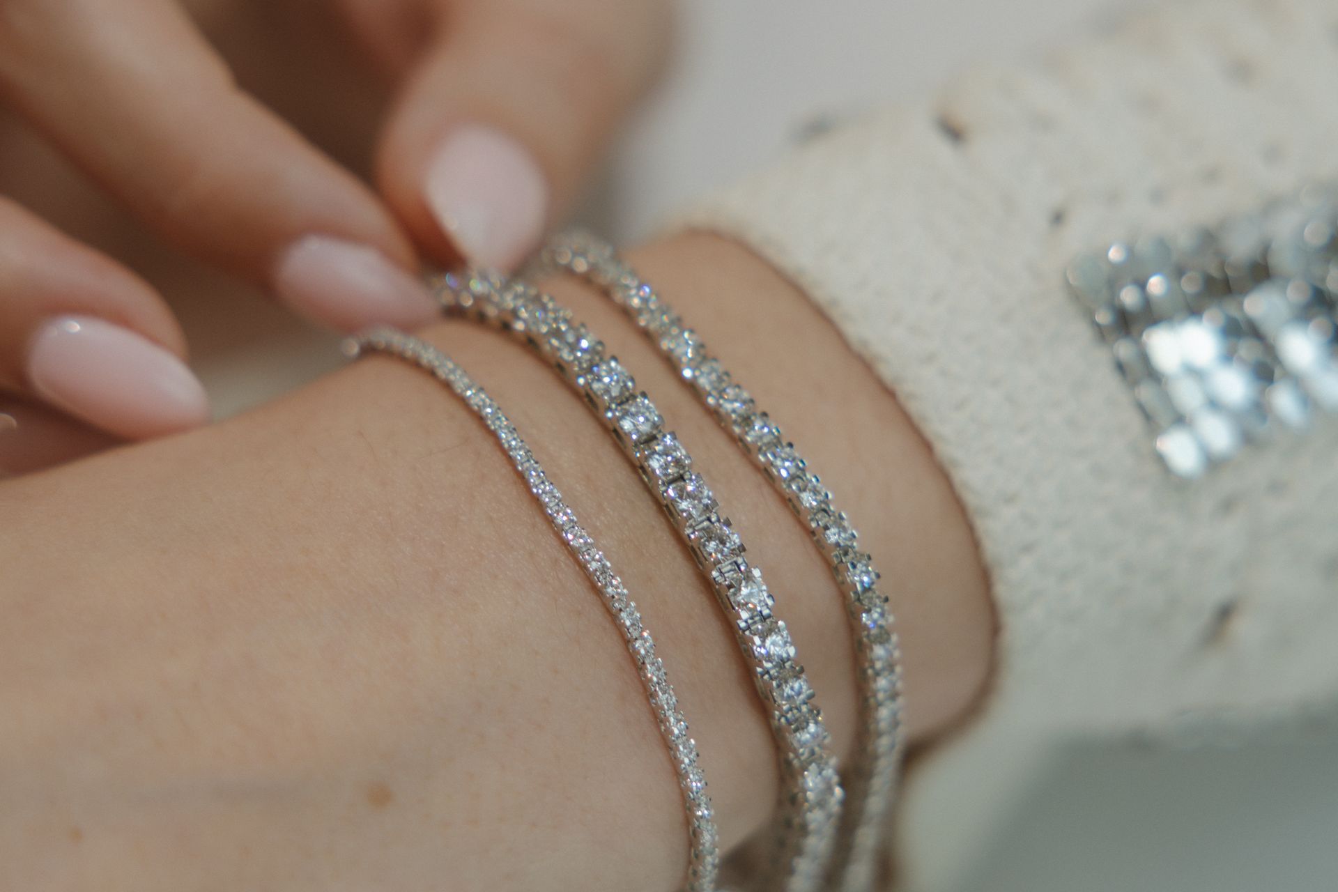 Close-up of a wrist with four silver bracelets, held by a hand with light pink nail polish, wearing a white sweater.