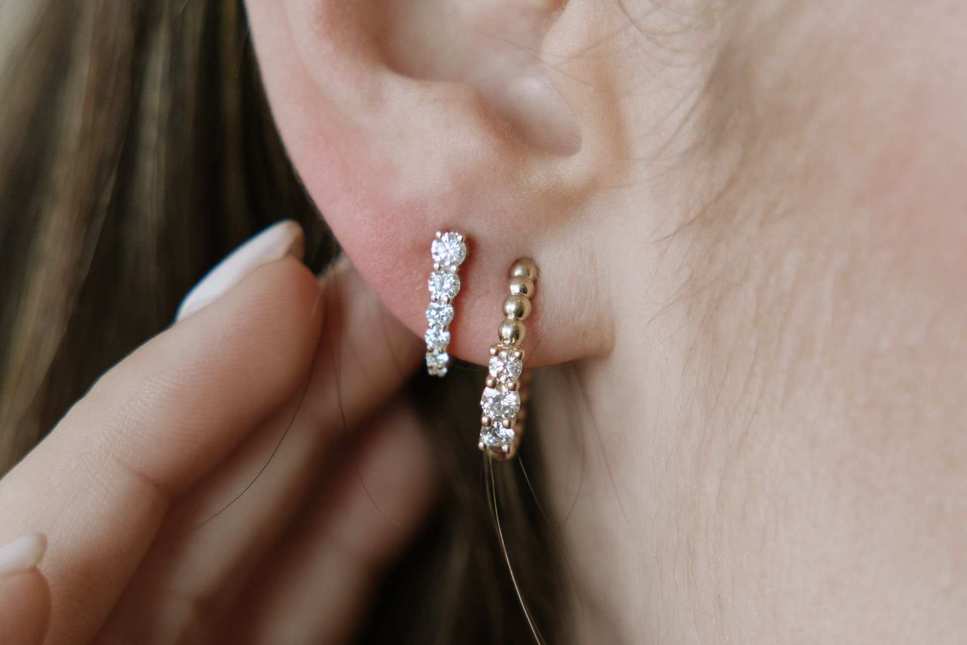 Close-up of ear with two sets of diamond earrings: one curved and one straight. A hand is adjusting one.